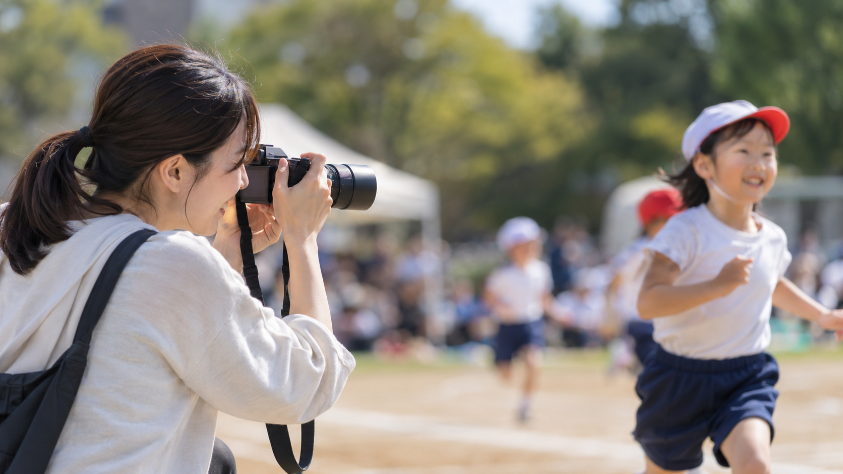 望遠レンズ付き小型ミラーレスカメラで運動会の子どもを撮影する日本人女性