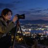 三脚にカメラを設置して夕暮れの街と港の夜景を撮影する日本人女性